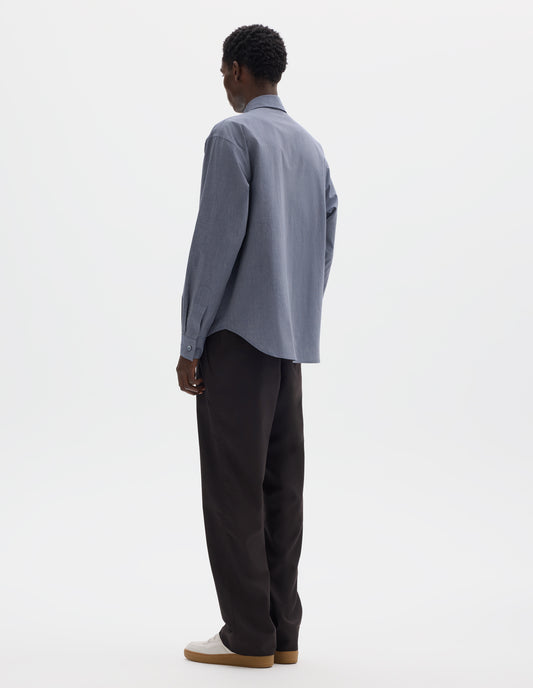 Full body image, back view of model wearing blue cotton shirt with mother of pearl buttons on cuffs and curved hem. Styled with dark brown trousers and beige nubuck trainers.