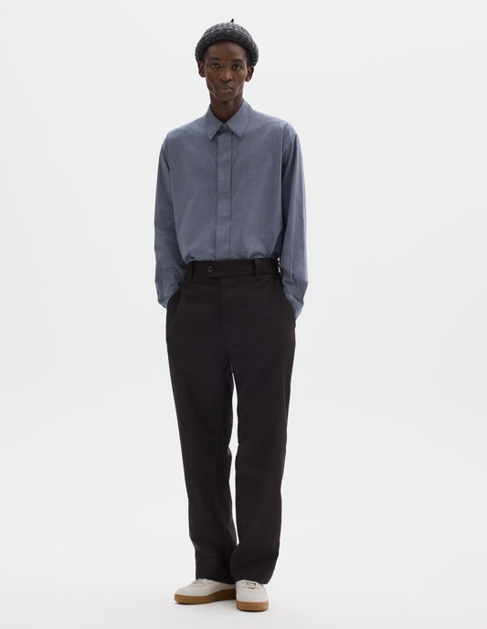 Full body image, front view of model wearing relaxed dark brown wool trousers with half-elasticated extended waistband, belt loops, horn button fastening and slant side pockets. Styled with grey beanie, blue shirt and beige nubuck trainers.
