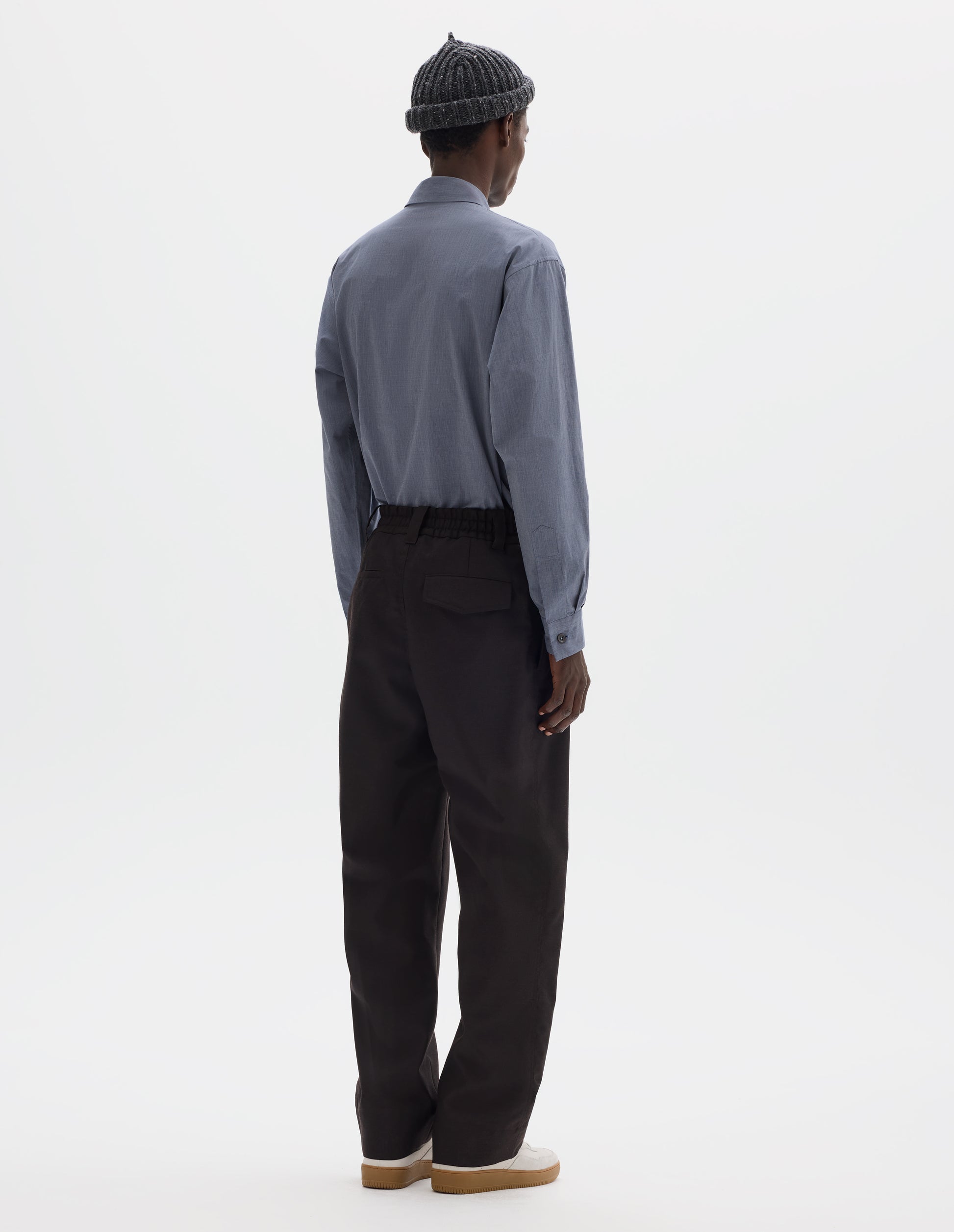 Full body image, back view of model wearing relaxed dark brown wool trousers with half-elasticated waist with belt loops, slant side pockets, and one welt back pocket and one flap back pocket with concealed button fastening. Styled with grey beanie, blue shirt and beige nubuck trainers.