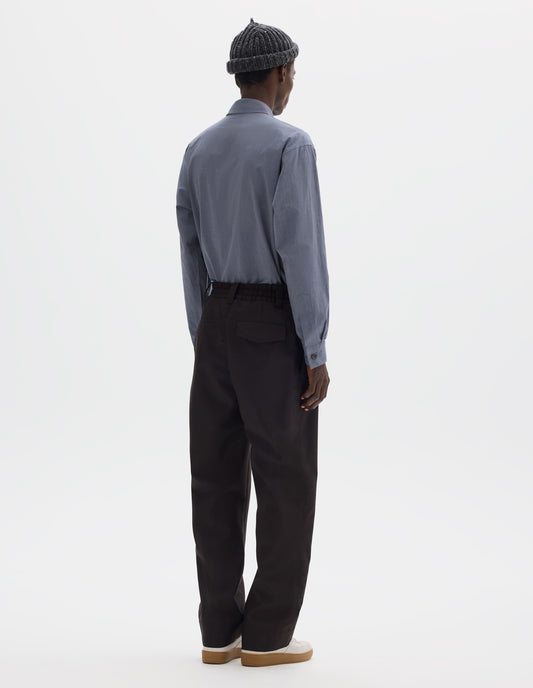 Full body image, back view of model wearing relaxed dark brown wool trousers with half-elasticated waist with belt loops, slant side pockets, and one welt back pocket and one flap back pocket with concealed button fastening. Styled with grey beanie, blue shirt and beige nubuck trainers.