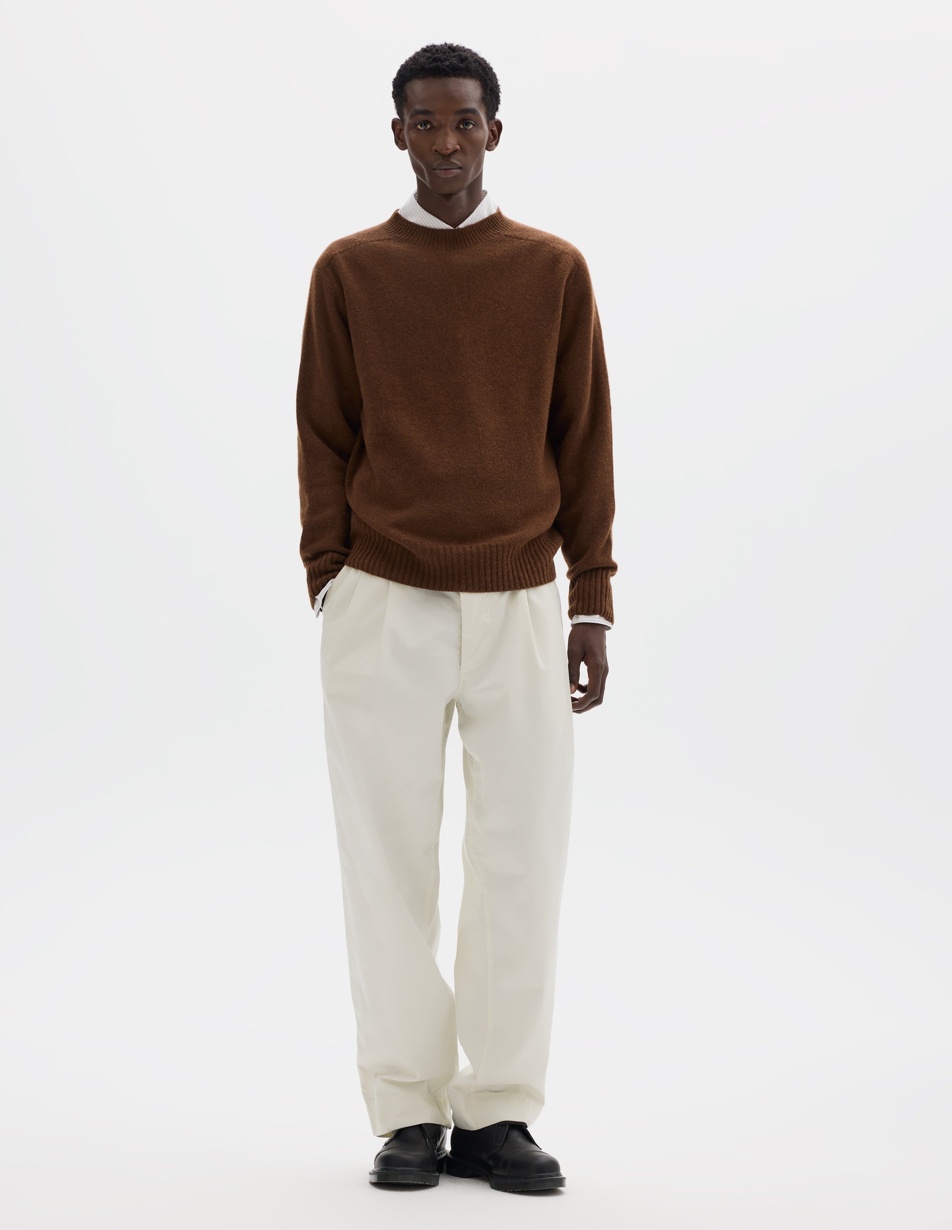 Full body image, front view of model wearing brown merino cashmere seamless jumper with ribbed crew neck, cuffs and hem. Styled over white shirt, and with white trousers and black leather shoes.