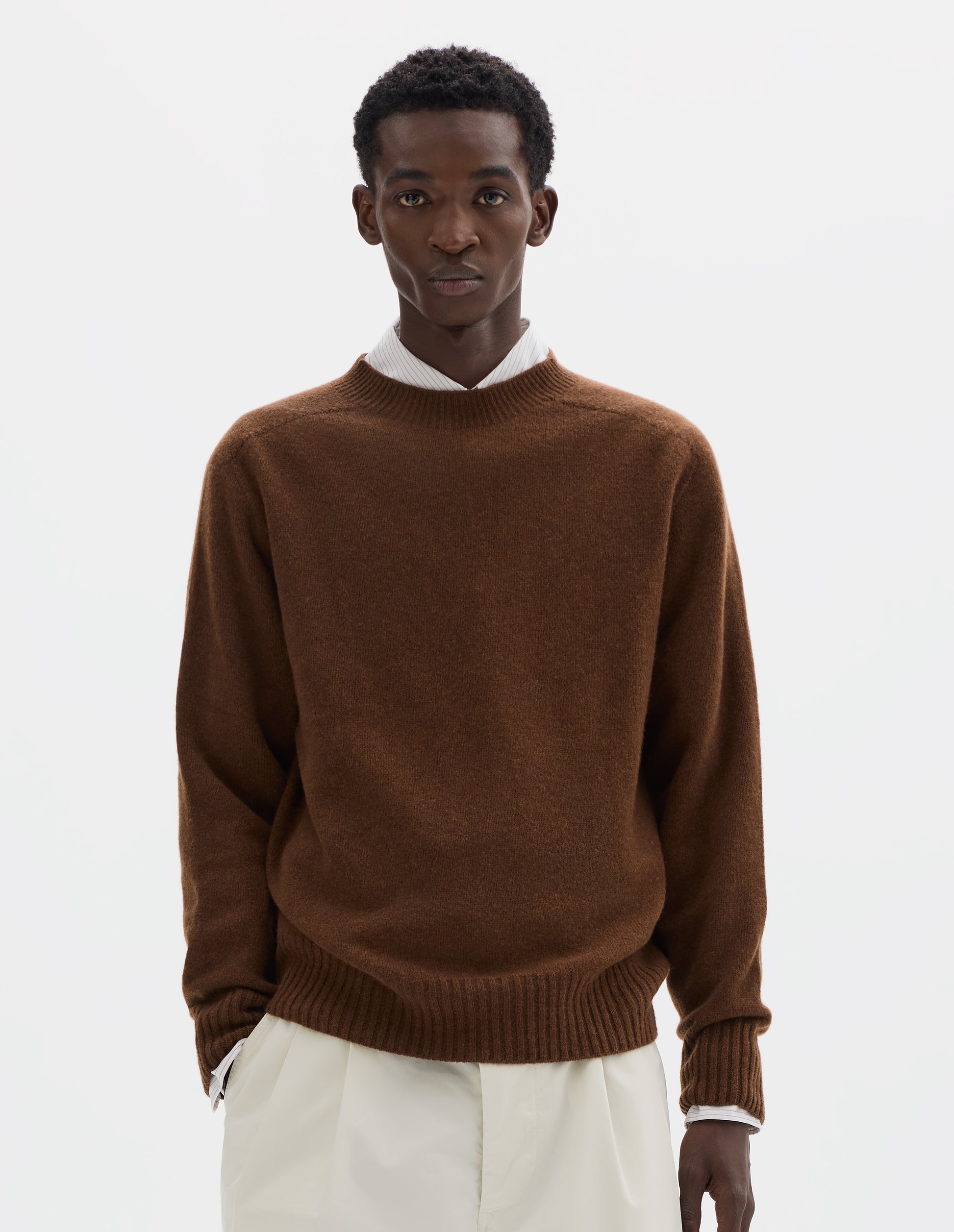 Cropped body image, front view of model wearing brown merino cashmere seamless jumper with ribbed crew neck, cuffs and hem. Styled over white shirt, and with white trousers.