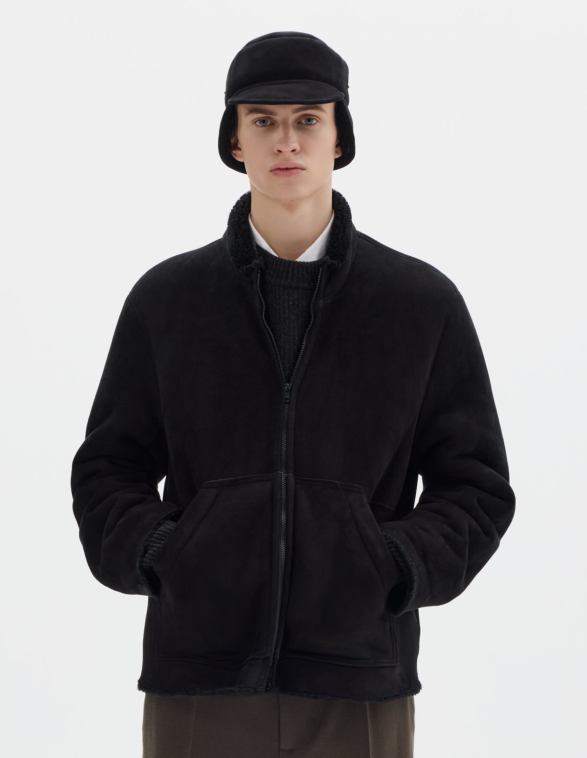 Cropped body image, front view of model wearing black relaxed sheepskin jacket with zip-through front, partial funnel neck with raw edge, slanted front half kangaroo pockets and raw edges on sleeves and hem. Styled over dark grey jumper and white shirt, and with matching shearling cap and khaki green trousers. 