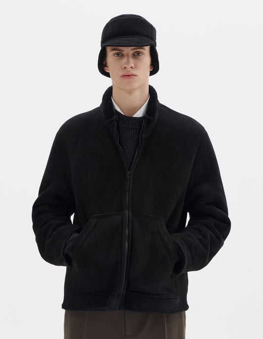 Cropped body image, front view of model wearing black relaxed sheepskin jacket with zip-through front, partial funnel neck with raw edge, slanted front half kangaroo pockets and raw edges on sleeves and hem. Styled over dark grey jumper and white shirt, and with matching shearling cap and khaki green trousers. 