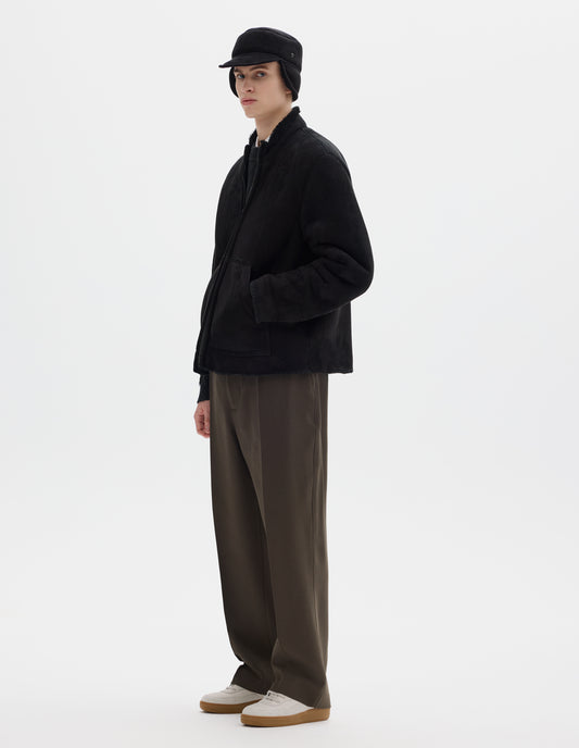Full body image, front view of model wearing black relaxed sheepskin jacket with zip-through front, partial funnel neck with raw edge, slanted front half kangaroo pockets and raw edges on sleeves and hem. Styled with matching shearling cap, khaki green trousers and beige nubuck trainers.
