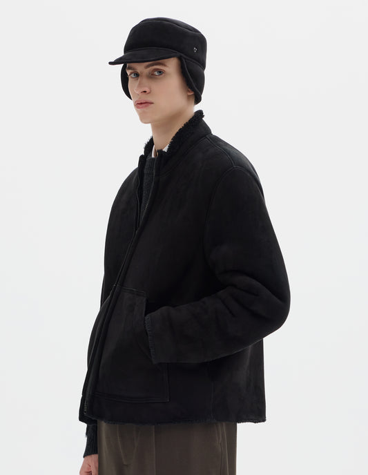 Cropped image, front view of male model wearing black shearling press stud cap with peak and black curly merino sheepskin ear flaps, worn down. Styled with matching black sheepskin coat and khaki green trousers.