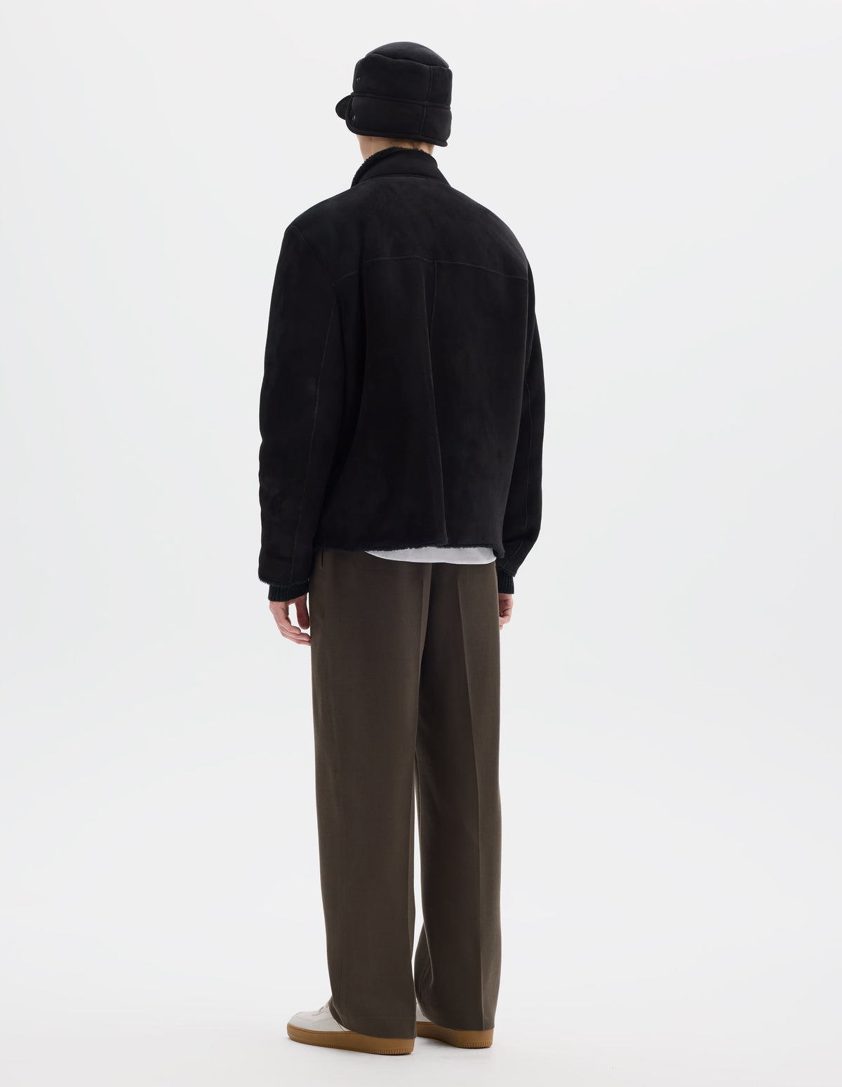 Full body image, back view of model wearing black relaxed sheepskin jacket with partial funnel neck, back yoke with a centre back seam and with raw edges on neck, sleeves and hem. Styled with matching shearling cap, khaki green trousers and beige nubuck trainers.