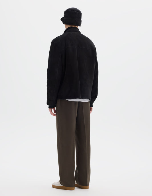 Full body image, back view of model wearing black relaxed sheepskin jacket with partial funnel neck, back yoke with a centre back seam and with raw edges on neck, sleeves and hem. Styled with matching shearling cap, khaki green trousers and beige nubuck trainers.