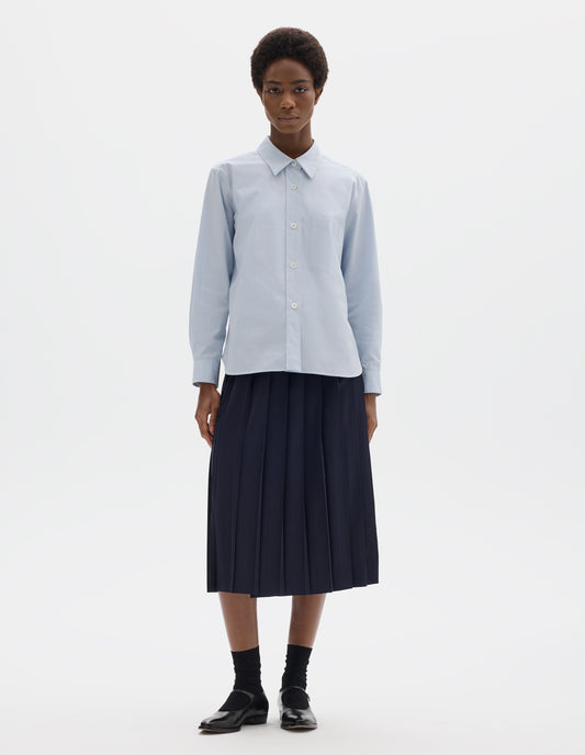 Full body image, front view of model wearing light blue cotton shirt with grey and beige stripes, mother of pearl buttons on raised placket, dropped patch pocket and curved hem. Styled with dark blue pleated skirt, black socks and black leather shoes.