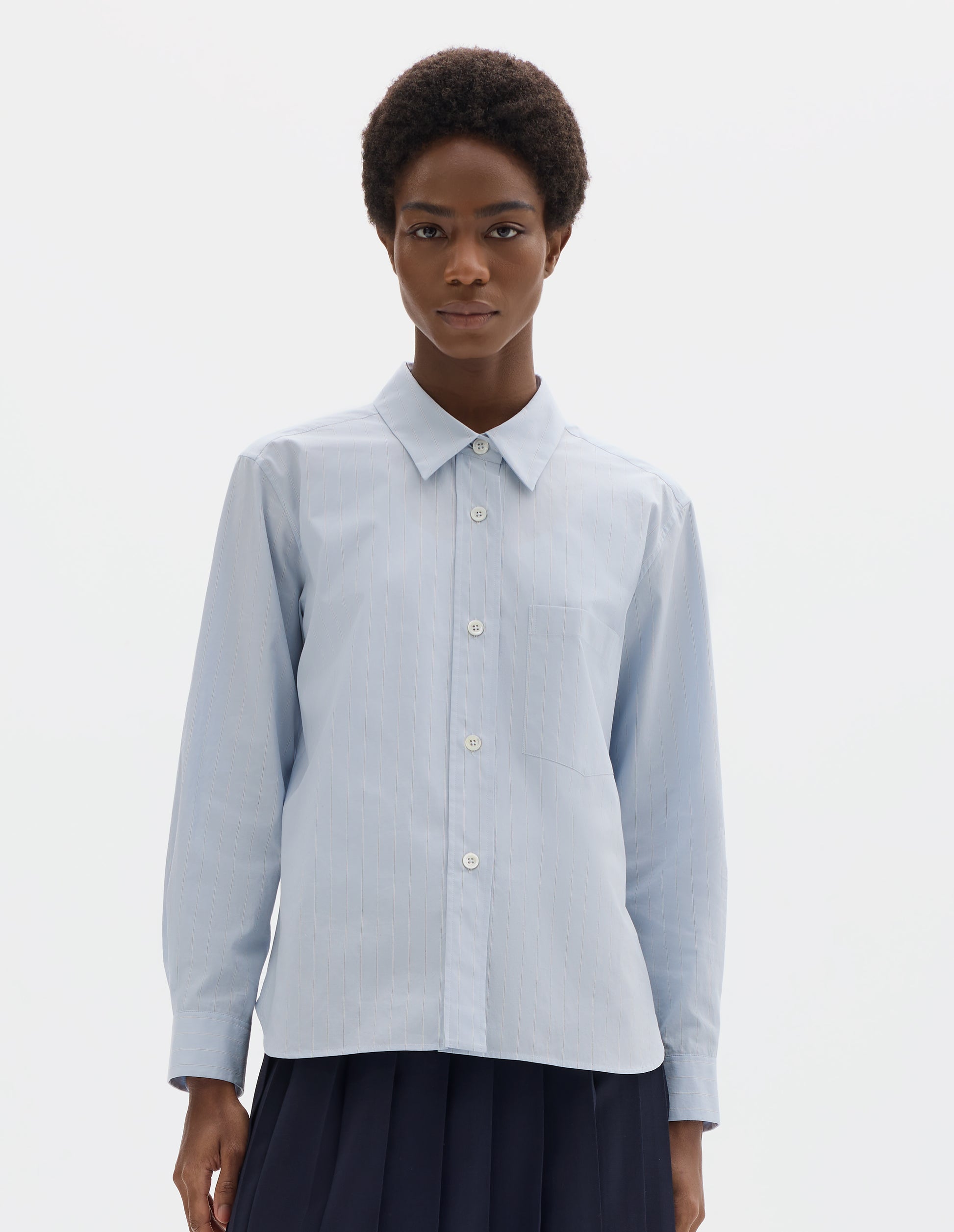Cropped body image, front view of model wearing light blue cotton shirt with grey and beige stripes, mother of pearl buttons on raised placket, chest patch pocket and curved hem. Styled with dark blue pleated skirt.