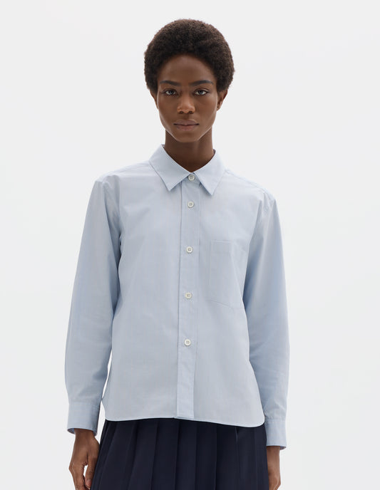 Cropped body image, front view of model wearing light blue cotton shirt with grey and beige stripes, mother of pearl buttons on raised placket, dropped patch pocket and curved hem. Styled with dark blue pleated skirt.