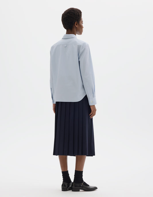 Full body image, back view of model wearing light blue cotton shirt with grey and beige stripes, hanger loop detail on back yoke, mother of pearl buttons on cuffs and curved hem. Styled with dark blue pleated skirt, black socks and black leather shoes.
