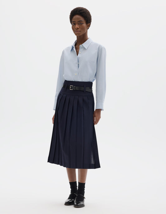 Full body image, front view of model wearing midi length dark blue wool full wrap skirt with stitched-on belt loops on dropped waistband, and concealed fastening with horn buttons. Styled with light blue shirt, black leather belt, black socks and black leather shoes.