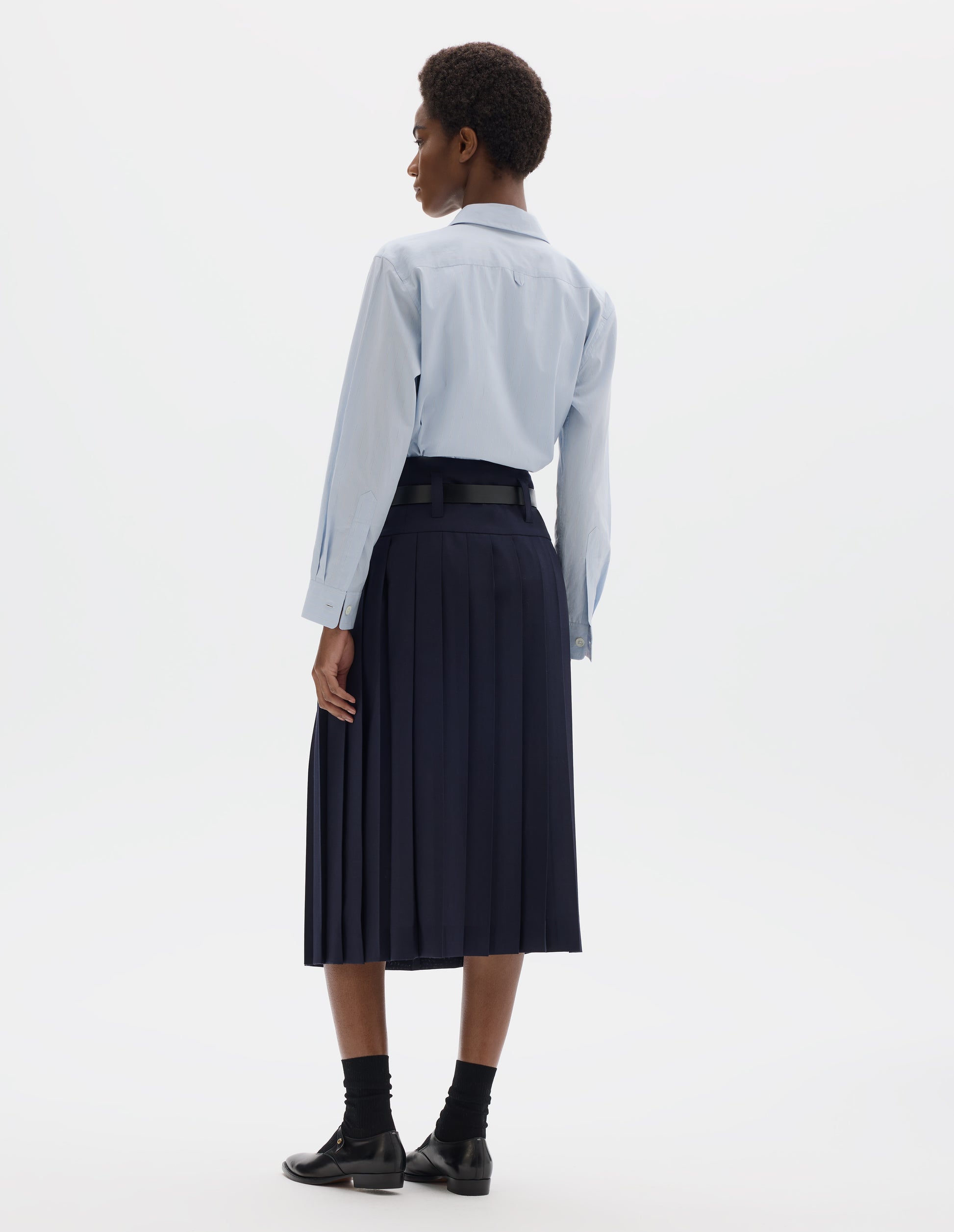 Full body image, back view of model wearing midi length dark blue wool full wrap skirt with stitched-on belt loops on dropped waistband.  Styled with light blue shirt, black leather belt, black socks and black leather shoes.