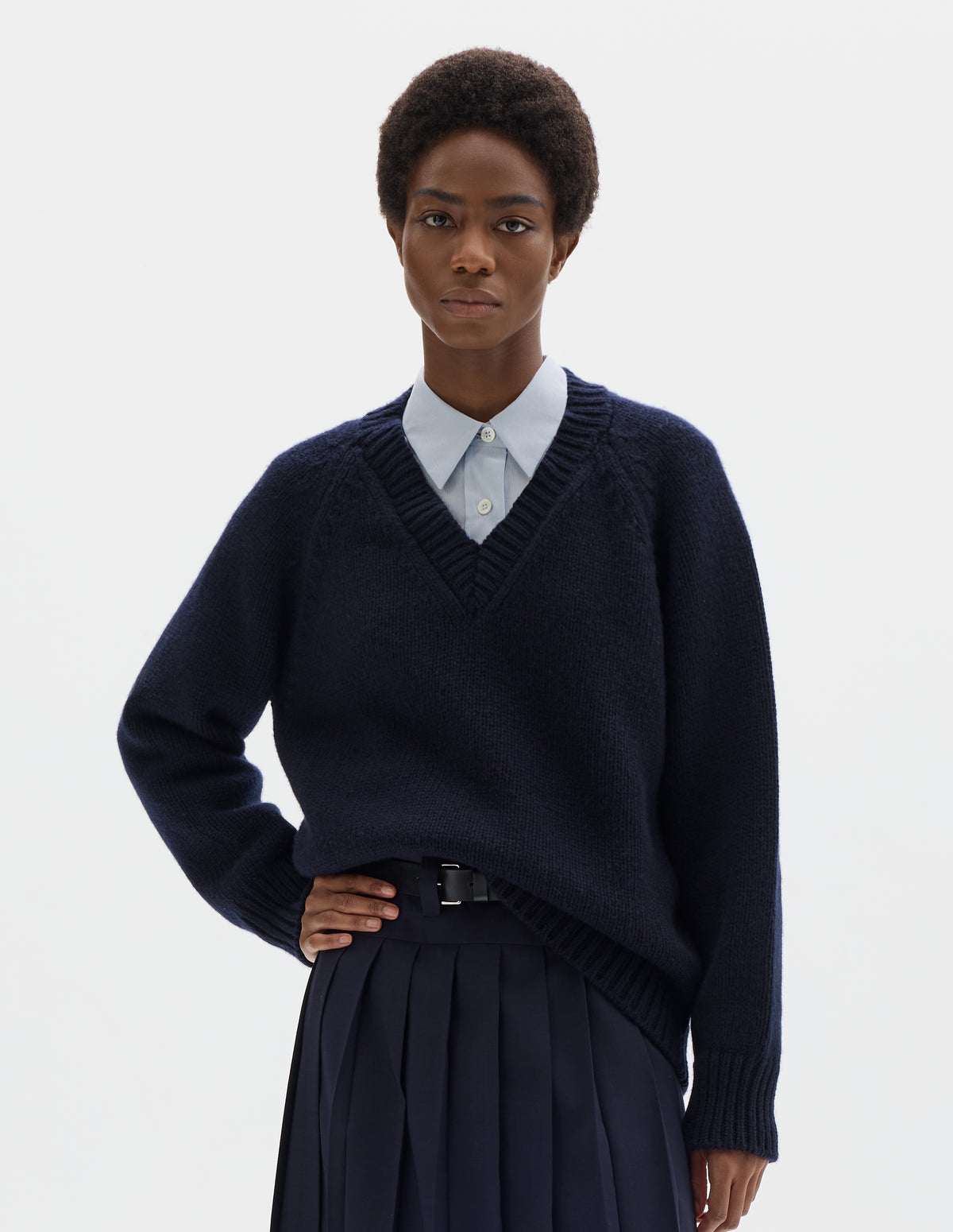 Cropped body image, front view of model wearing oversized dark blue cashmere wool jumper with V neck, raglan sleeves and fully fashioned arm holes, and with ribbed neck, cuffs and hem. Styled over light blue shirt, and with dark blue skirt with black leather belt.