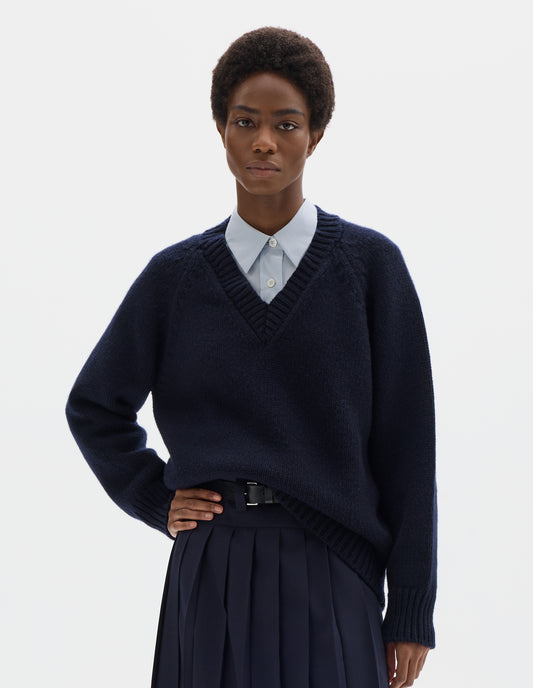 Cropped body image, front view of model wearing oversized dark blue cashmere wool jumper with V neck, raglan sleeves and fully fashioned arm holes, and with ribbed neck, cuffs and hem. Styled over light blue shirt, and with dark blue skirt with black leather belt.