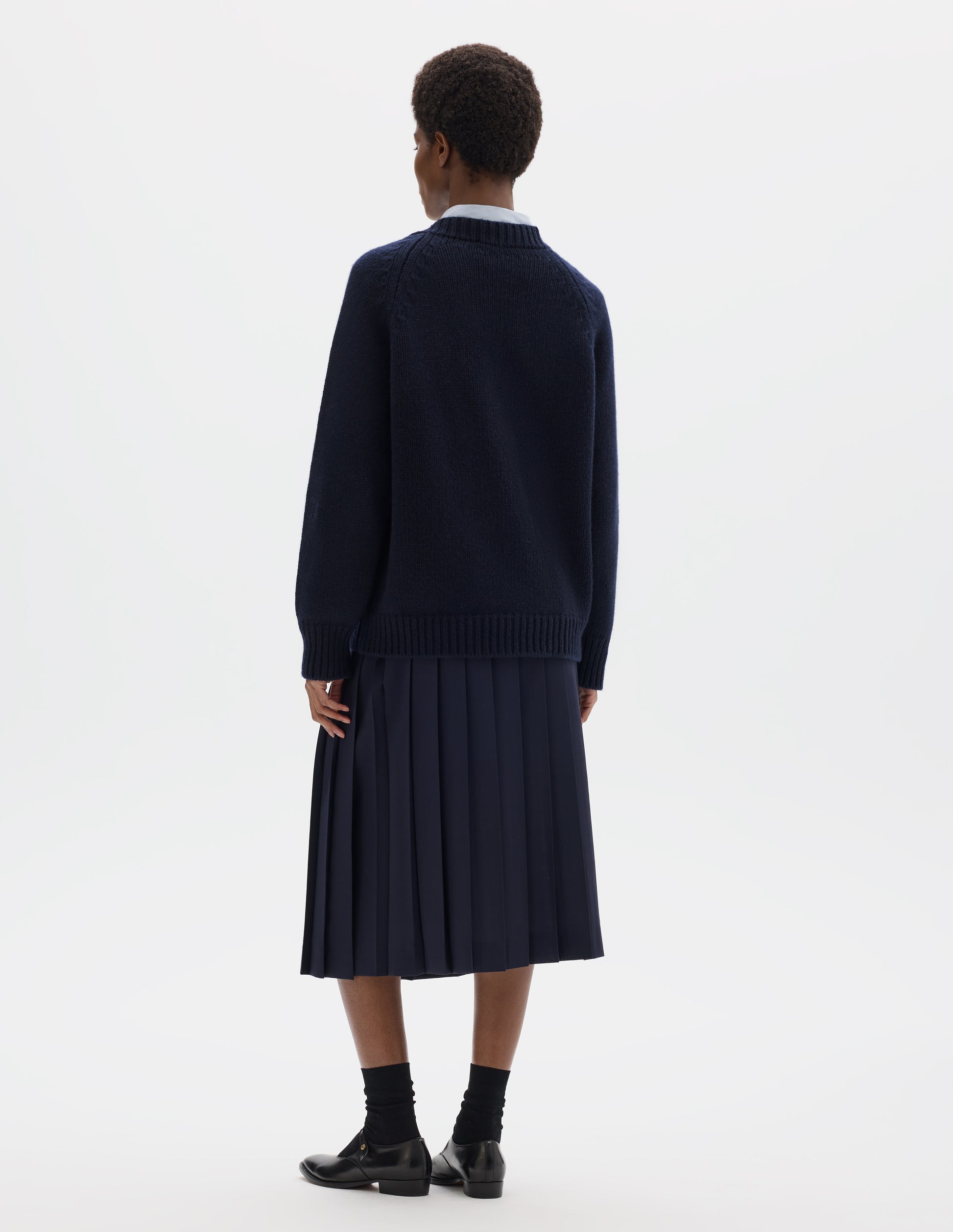 Full body image, back view of model wearing oversized dark blue cashmere wool jumper with raglan sleeves and fully fashioned arm holes, and with ribbed neck, cuffs and hem. Styled over light blue shirt and with dark blue skirt, black socks and black leather shoes.