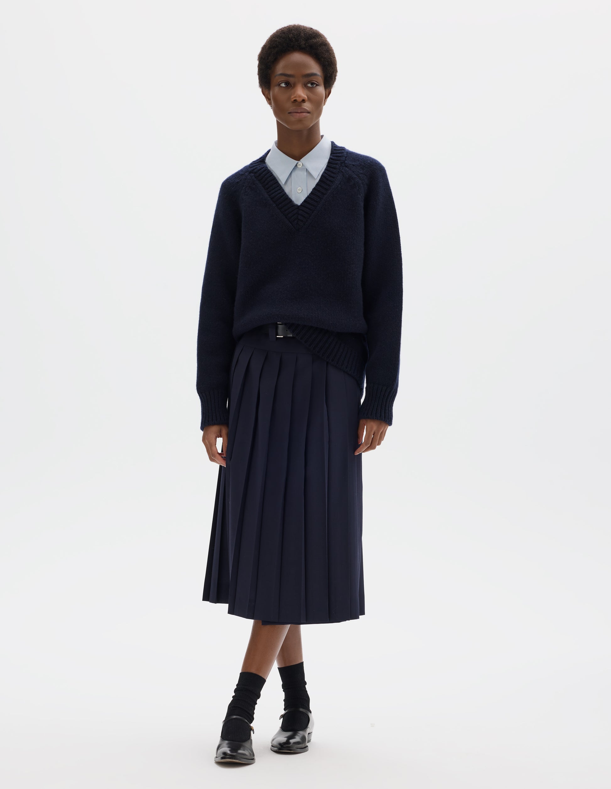 Full body image, front view of model wearing oversized dark blue cashmere wool jumper with V neck, raglan sleeves and fully fashioned arm holes, and with ribbed neck, cuffs and hem. Styled over light blue shirt and with dark blue skirt with black leather belt, black socks and black leather shoes.