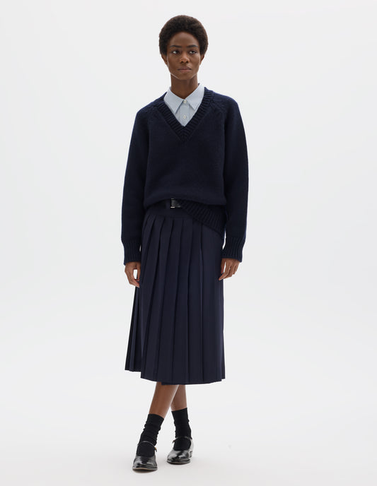Full body image, front view of model wearing oversized dark blue cashmere wool jumper with V neck, raglan sleeves and fully fashioned arm holes, and with ribbed neck, cuffs and hem. Styled over light blue shirt and with dark blue skirt with black leather belt, black socks and black leather shoes.