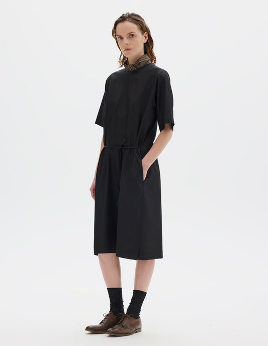Full body image, front view of model wearing black cotton blend midi A line dress with bias cut binding neckline, deep hem sleeves, thin belt in self fabric, side seam pockets, and deep vent split hem. Styled with khaki green silk scarf, black socks and brown leather brogues.