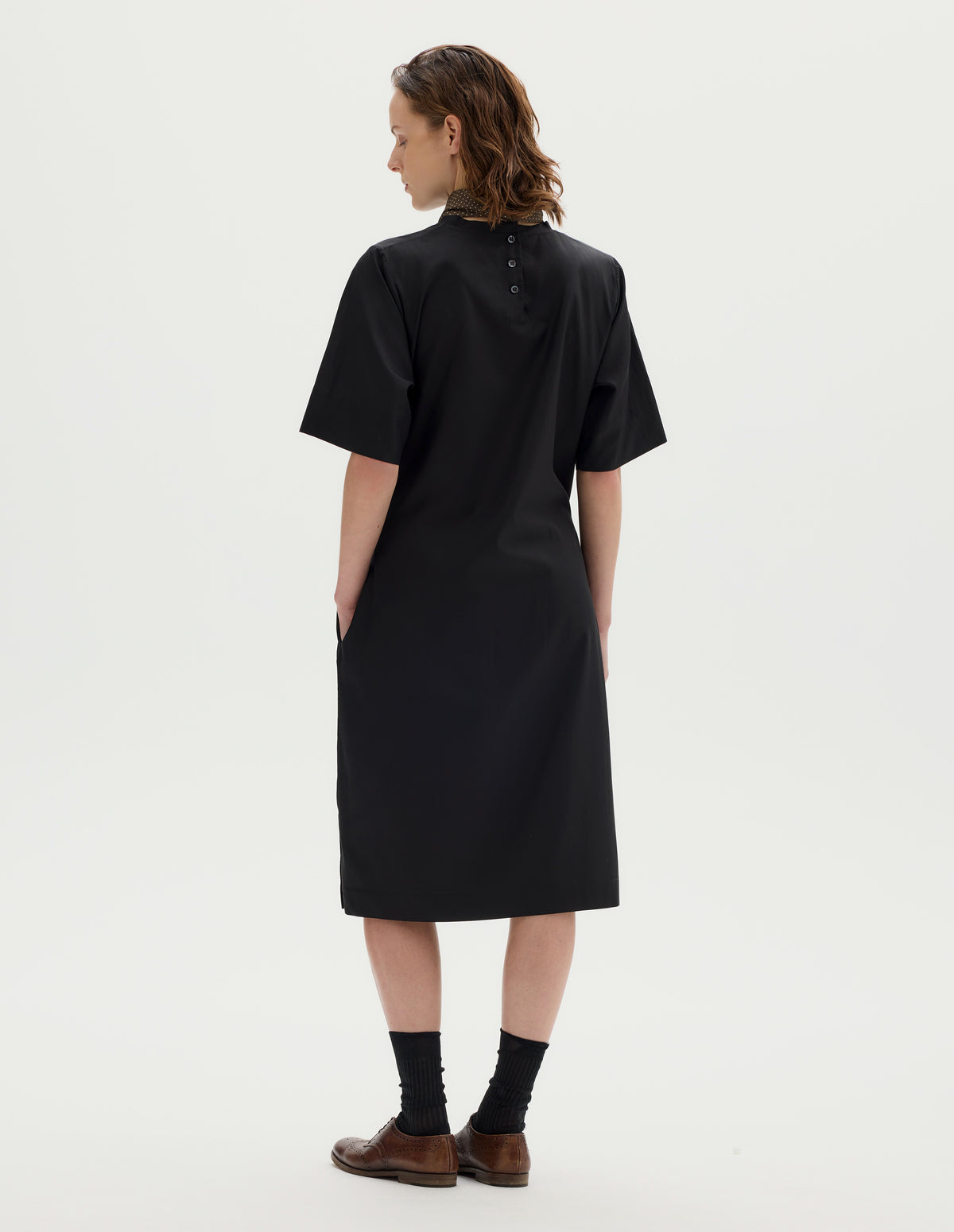 Full body image, back view of model wearing black cotton blend midi A line dress with cut bias cut binding neckline, three mother of pearl buttons at centre, deep hem sleeves, side seam pocekts and deep vent split hem. Styled with khaki green silk scarf, black socks and brown leather brogues.