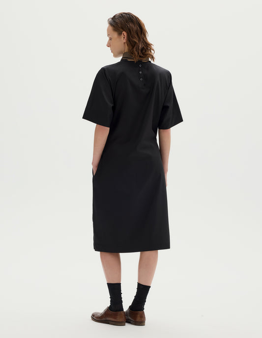 Full body image, back view of model wearing black cotton blend midi A line dress with cut bias cut binding neckline, three mother of pearl buttons at centre, deep hem sleeves, side seam pocekts and deep vent split hem. Styled with khaki green silk scarf, black socks and brown leather brogues.