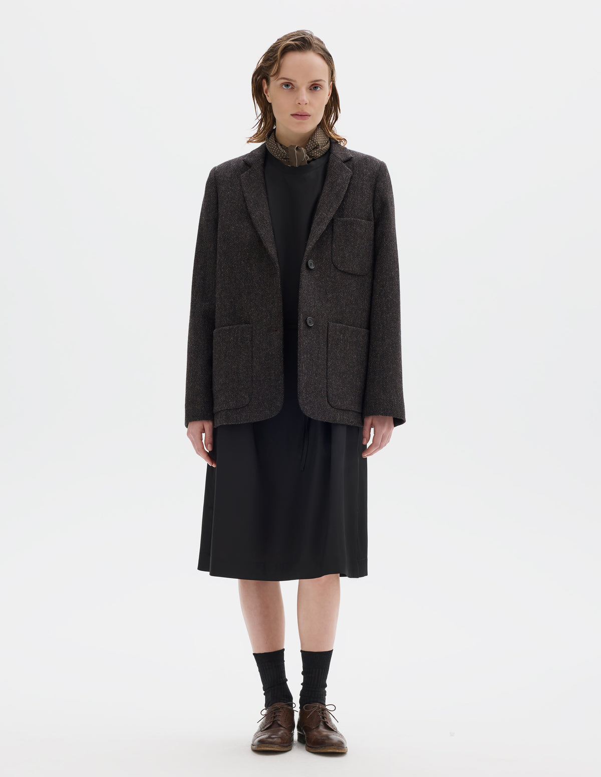 Full body image, front view of model wearing brown and black herringbone wool blazer with narrow collar and horn button-through revere, three bluff patch pockets, rounded details on hem and pockets. Styled over black dress and with khaki green silk scarf, black socks and brown leather brogues.