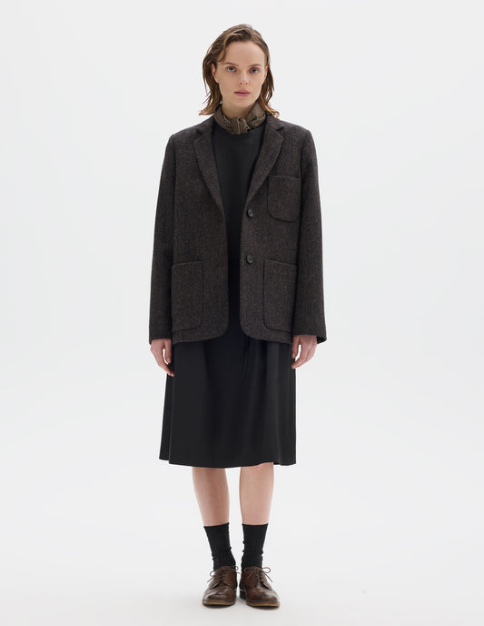 Full body image, front view of model wearing brown and black herringbone wool blazer with narrow collar and horn button-through revere, three bluff patch pockets, rounded details on hem and pockets. Styled over black dress and with khaki green silk scarf, black socks and brown leather brogues.