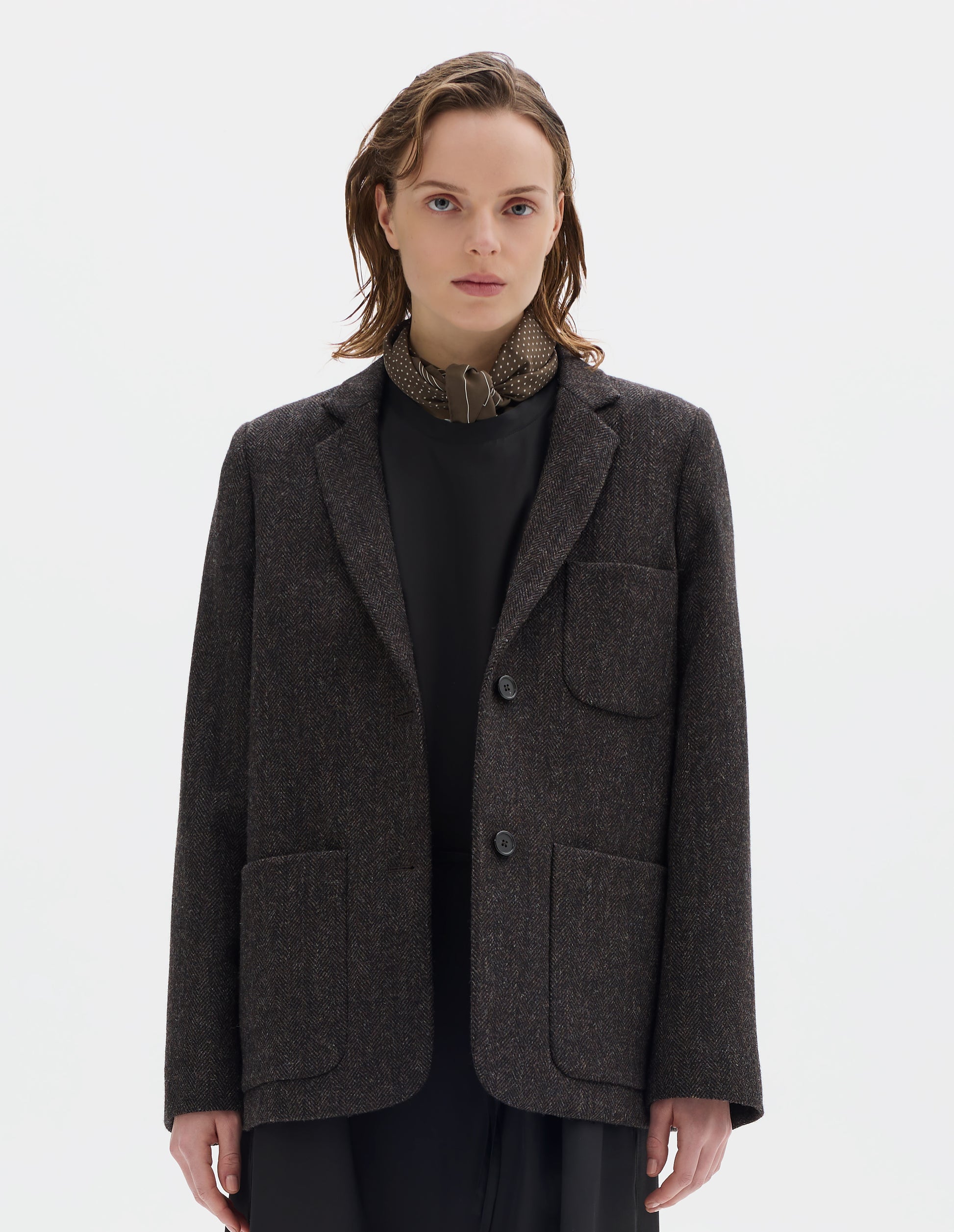 Cropped body image, front view of model wearing brown and black herringbone wool blazer with narrow collar and horn button-through revere, three bluff patch pockets, rounded details on hem and pockets. Styled over black dress and with khaki green silk scarf.