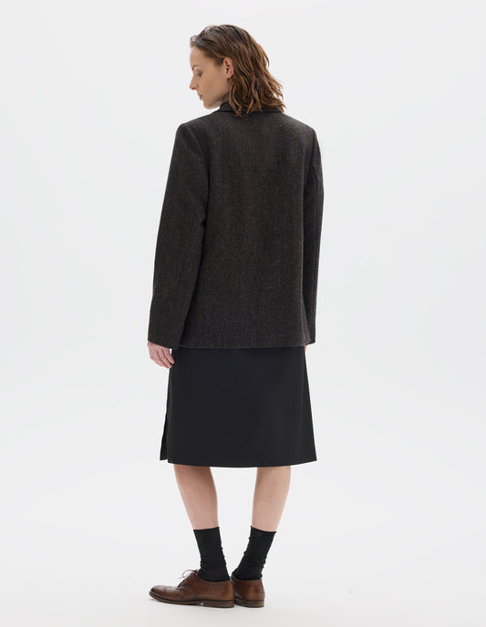 Full body image, back view of model wearing relaxed brown and black herringbone wool blazer with narrow collar. Styled over black dress and with black socks and brown leather brogues.