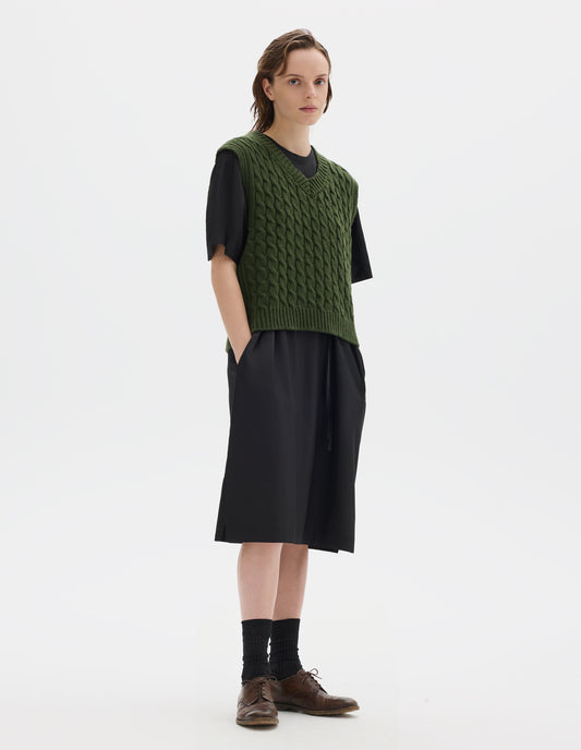 Full body image, front view of model wearing  relaxed green wool slipover with V neck, front cable design and ribbed neck, armholes, side seams and hem. Styled over black dress, and with black socks and brown leather brogues.