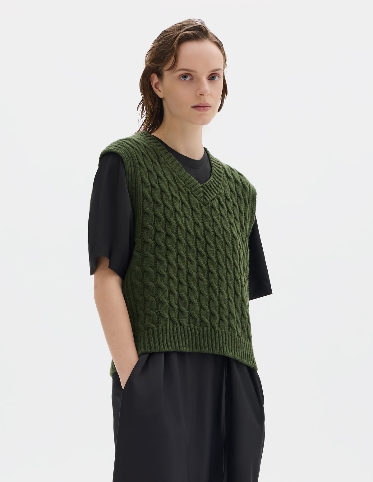 Cropped body image, front view of model wearing relaxed green wool slipover with V neck, front cable design and ribbed neck, armholes, side seams and hem. Styled over black dress.