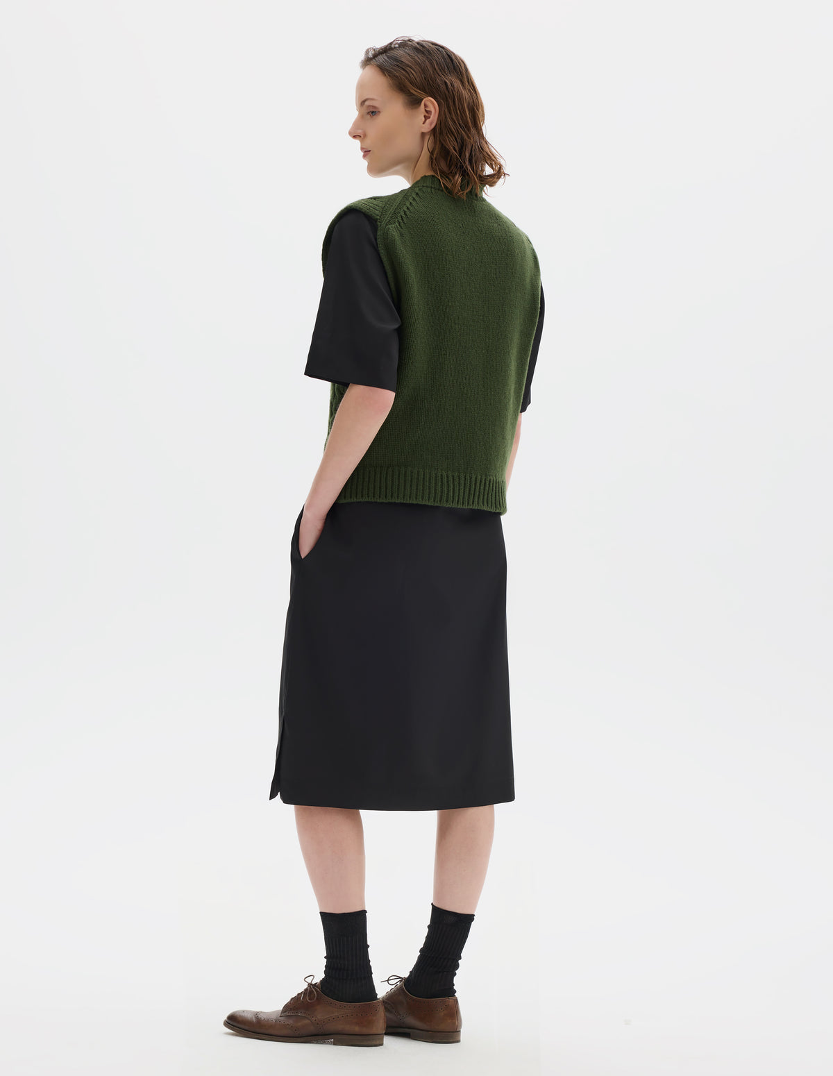 Full body image, back view of model wearing relaxed green wool slipover with plain knitted back and ribbed neck, armholes, side seams and hem. Styled over black dress, and with black socks and brown leather brogues.