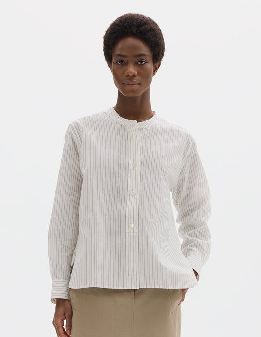 Cropped body image, front view of model wearing white collarless cotton and silk shirt with grey stripes, mother of pearl buttons on raised placket with box stitch detailing and curved hem. Styled with beige skirt.