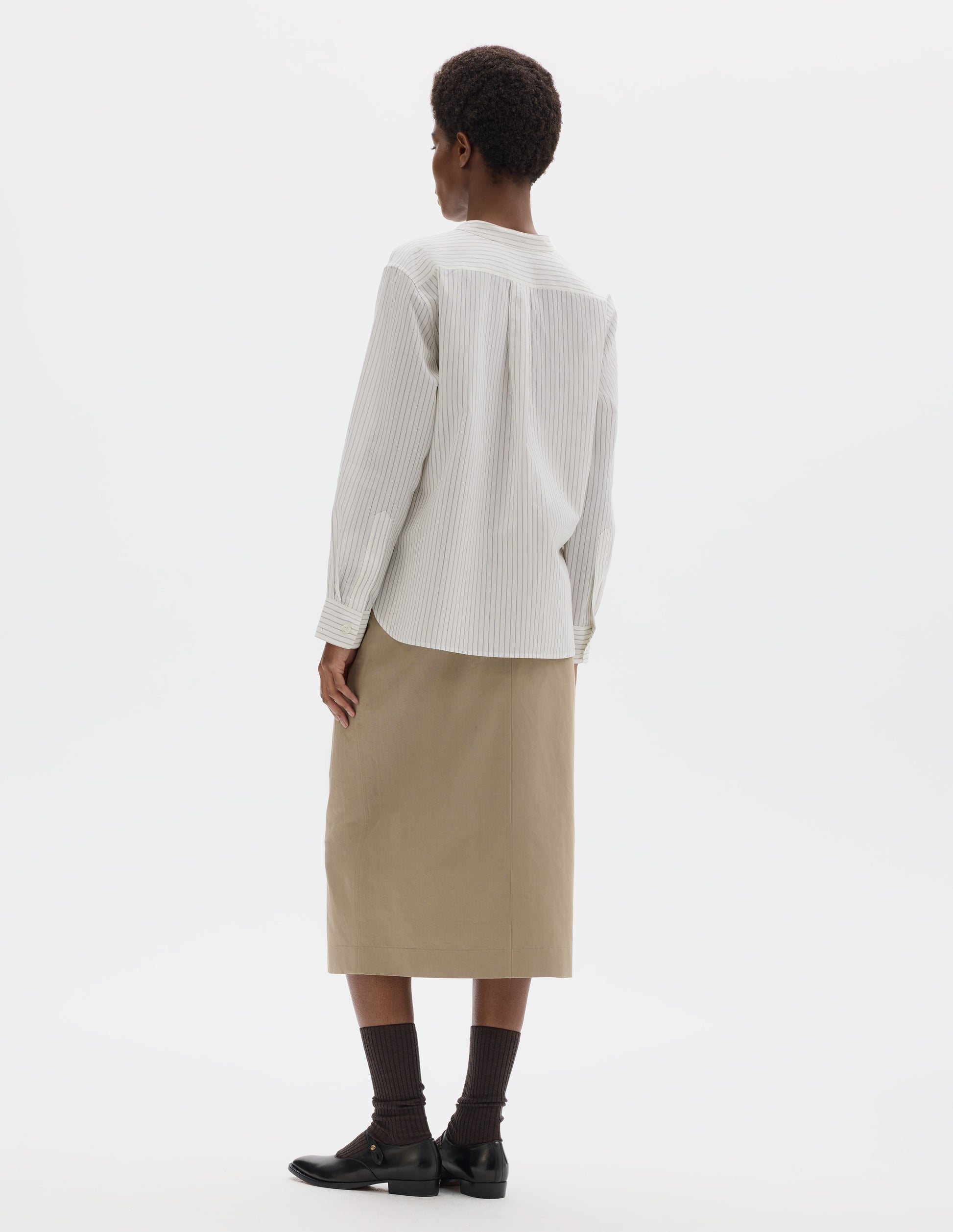 Full body image, back view of model wearing white collarless cotton and silk shirt with grey stripes, buttonhole detailing on collar band and box pleat on back yoke, mother of pearl buttons cuffs, elongated curved back hem. Styled with beige skirt, dark brown socks and black leather shoes.