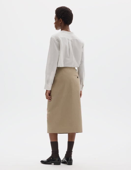 Full body image, back view of model wearing beige cotton linen skirt with horn button side cinches, horn button-through back welt pocket, and with stitch-through hem. Styled with white striped shirt, black socks and black leather shoes.