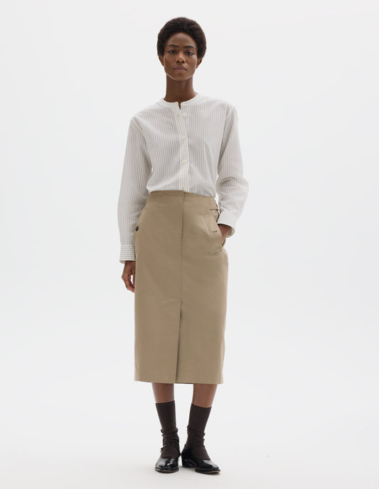 Full body image, front view of model wearing beige cotton linen skirt with concealed zip fly and hook and bar fastening, waist cinch detail, horn button-through slanted welt pockets, centre front vent split stitch-through hem. Styled with white striped shirt, black socks and black leather shoes.