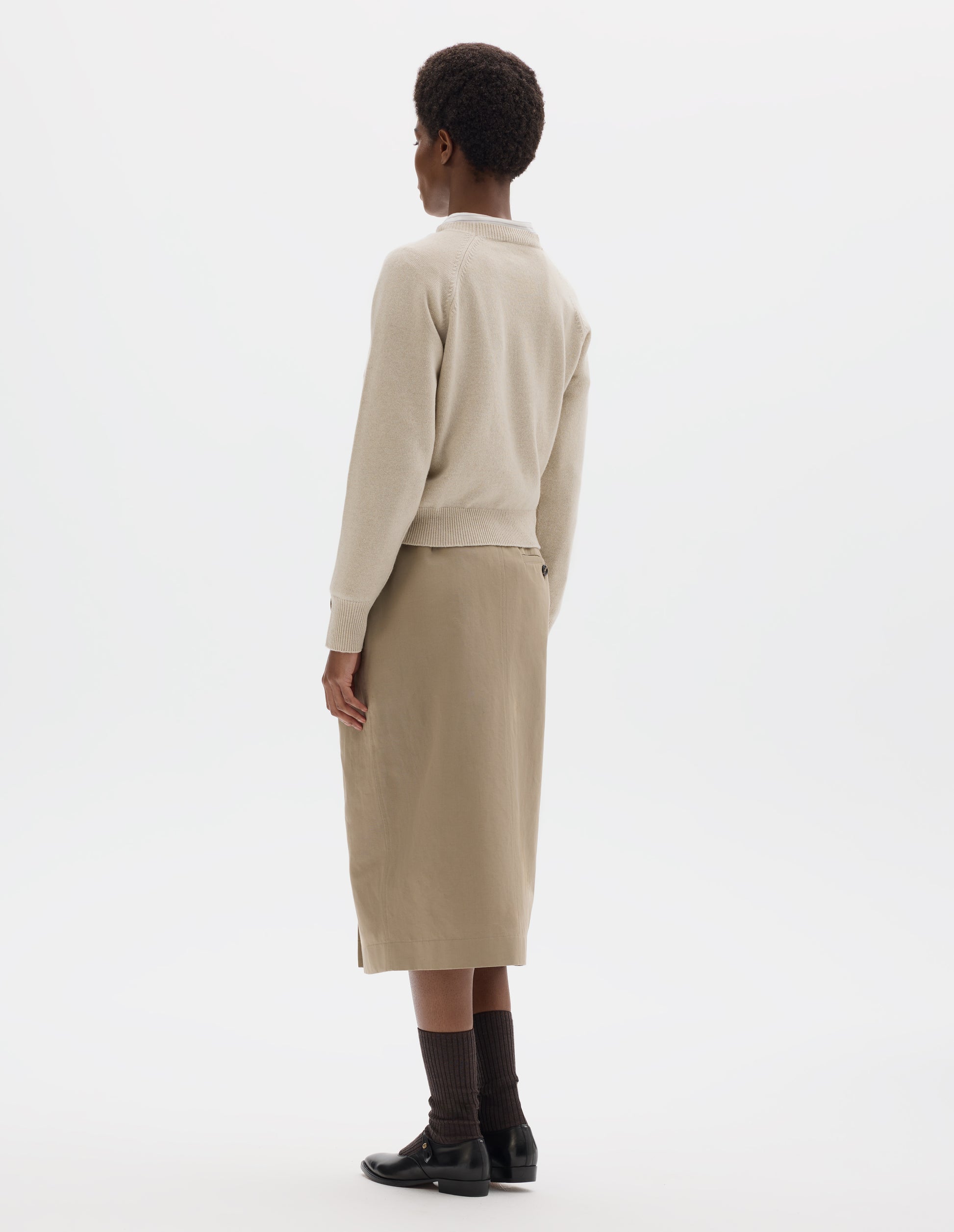 Full body image, back view of model wearing light beige merino cashmere jumper with fully fashioned raglan sleeves with ribbed cuffs and ribbed hem. Styled with beige skirt, dark brown socks and black leather shoes.