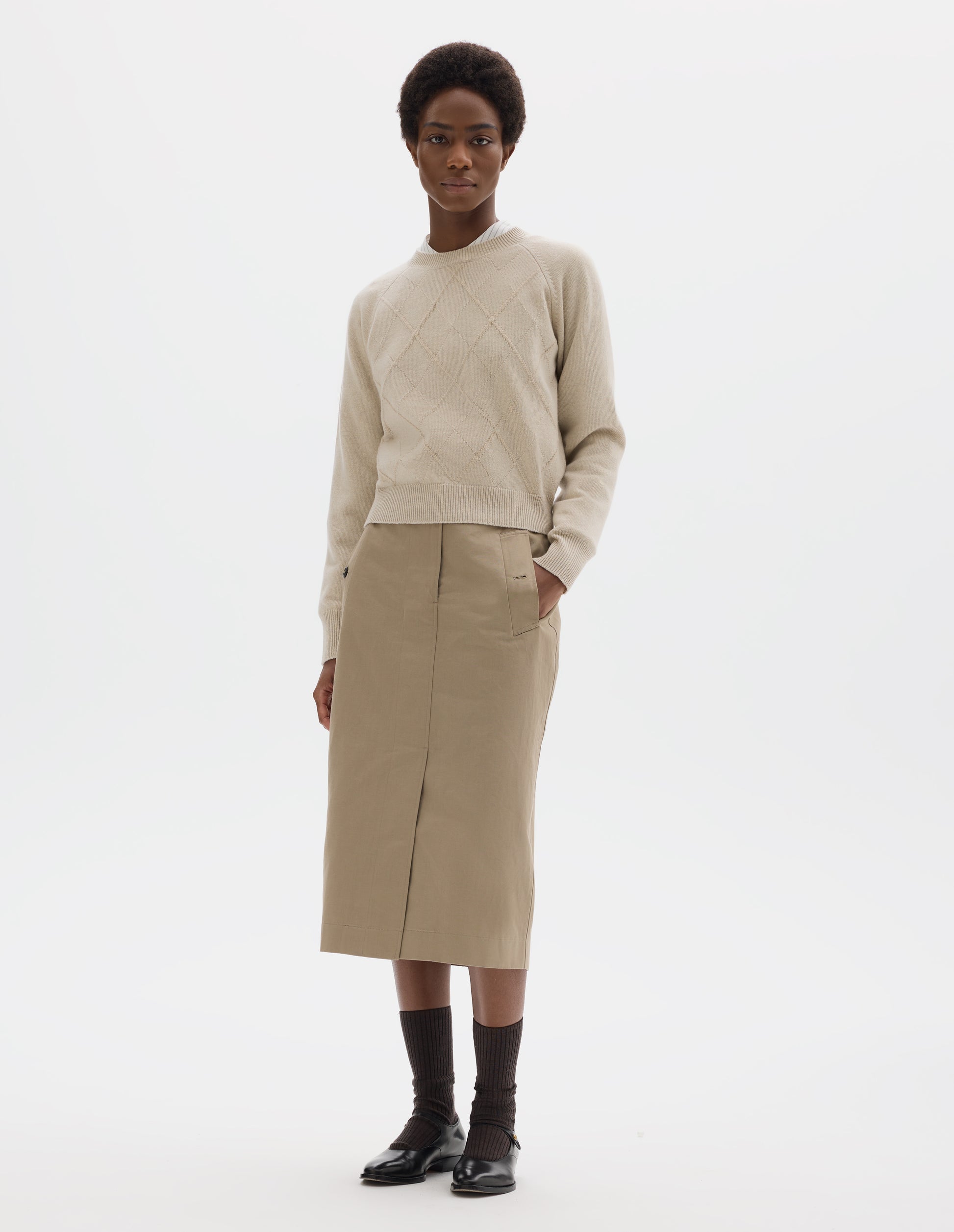 Full body image, front view of model wearing light beige merino cashmere jumper with textural argyle design, fully fashioned raglan sleeves with ribbed cuffs and ribbed hem. Styled over white shirt, and with beige skirt, dark brown socks and black leather shoes.