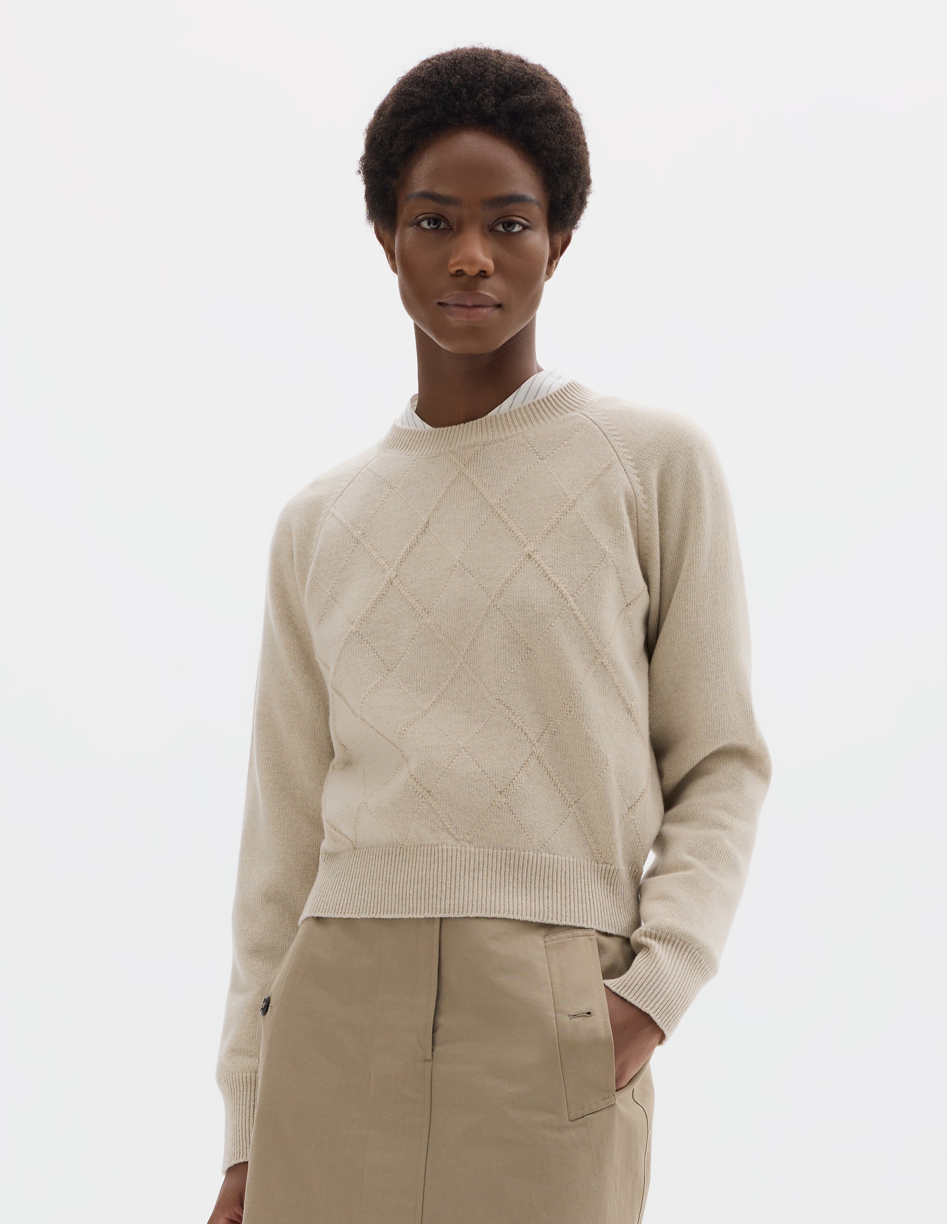 Cropped body image, front view of model wearing light beige merino cashmere jumper with textural argyle design, fully fashioned raglan sleeves with ribbed cuffs and ribbed hem. Styled over white shirt, and with beige skirt