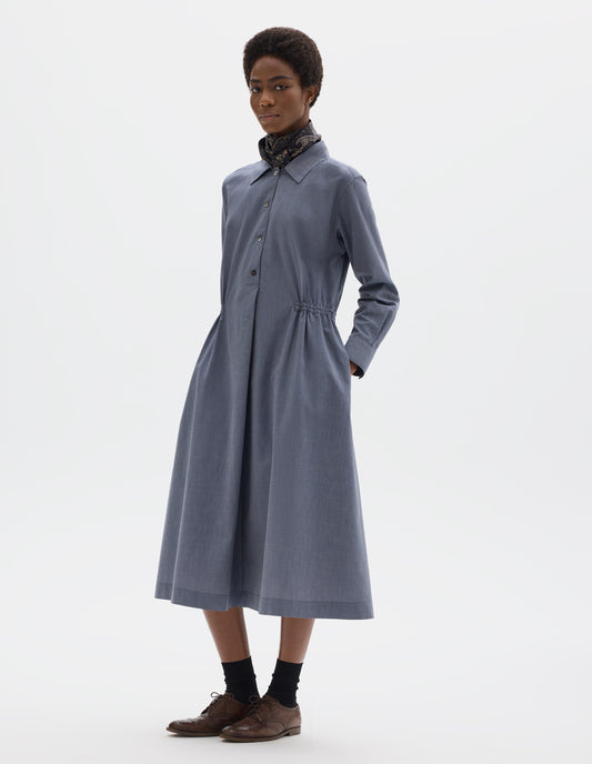 Full body image, front view of model wearing relaxed blue cotton midi dress with collar, mother of pearls buttons on placket, long shirt sleeves, side pockets and externally applied channel at waist. Styled with dark blue paisley neck scarf, black socks and brown leather brogues.