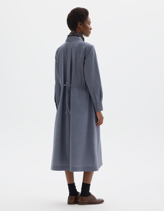 Full body image, back view of model wearing relaxed blue cotton midi dress with collar, pleat on narrow yoke on back, shirt sleeves with mother of pearl buttons on cuffs, externally applied channel at waist with double tie. Styled with dark blue paisley neck scarf, black socks and brown leather brogues.