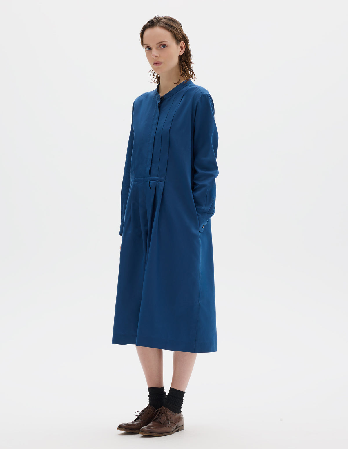 Full body image, front view of model wearing blue cotton silk collarless midi dress with concealed half placket on shirt front with single mother of pearl button fastening, shirt sleeves with mother of pearl buttons on narrow cuffs, side seam pockets, deep stitch-through pleats on front bib and skirt, and split hems. Styled with black socks and brown leather brogues.