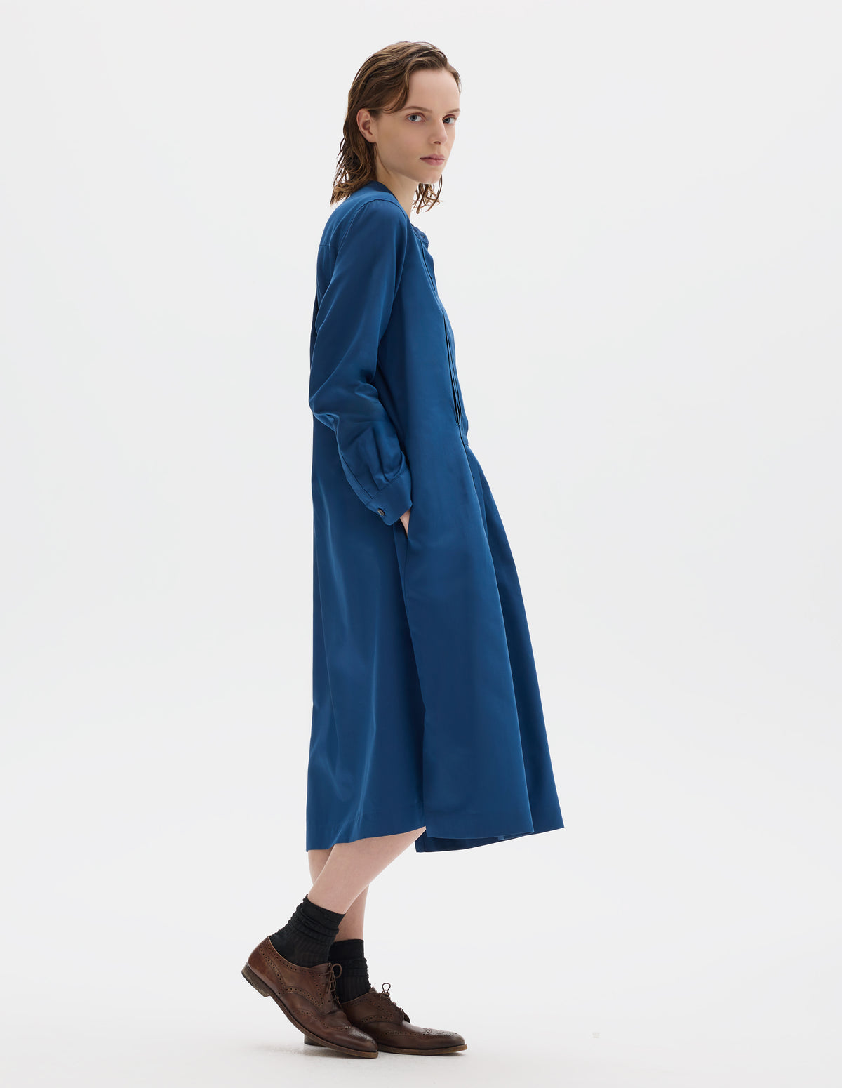 Full body image, side view of model wearing blue cotton silk collarless midi dress with concealed half placket on shirt front with single mother of pearl button fastening, shirt sleeves with mother of pearl buttons on narrow cuffs, side seam pockets, deep stitch-through pleats on front bib and skirt, and split hems. Styled with black socks and brown leather brogues.
