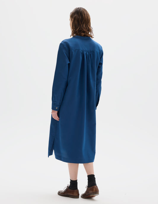 Full body image, back view of model wearing blue cotton silk collarless midi dress with wide gathering at back yoke, shirt sleeves with mother of pearl buttons on narrow cuffs, side seam pockets and  split hems. Styled with black socks and brown leather brogues.
