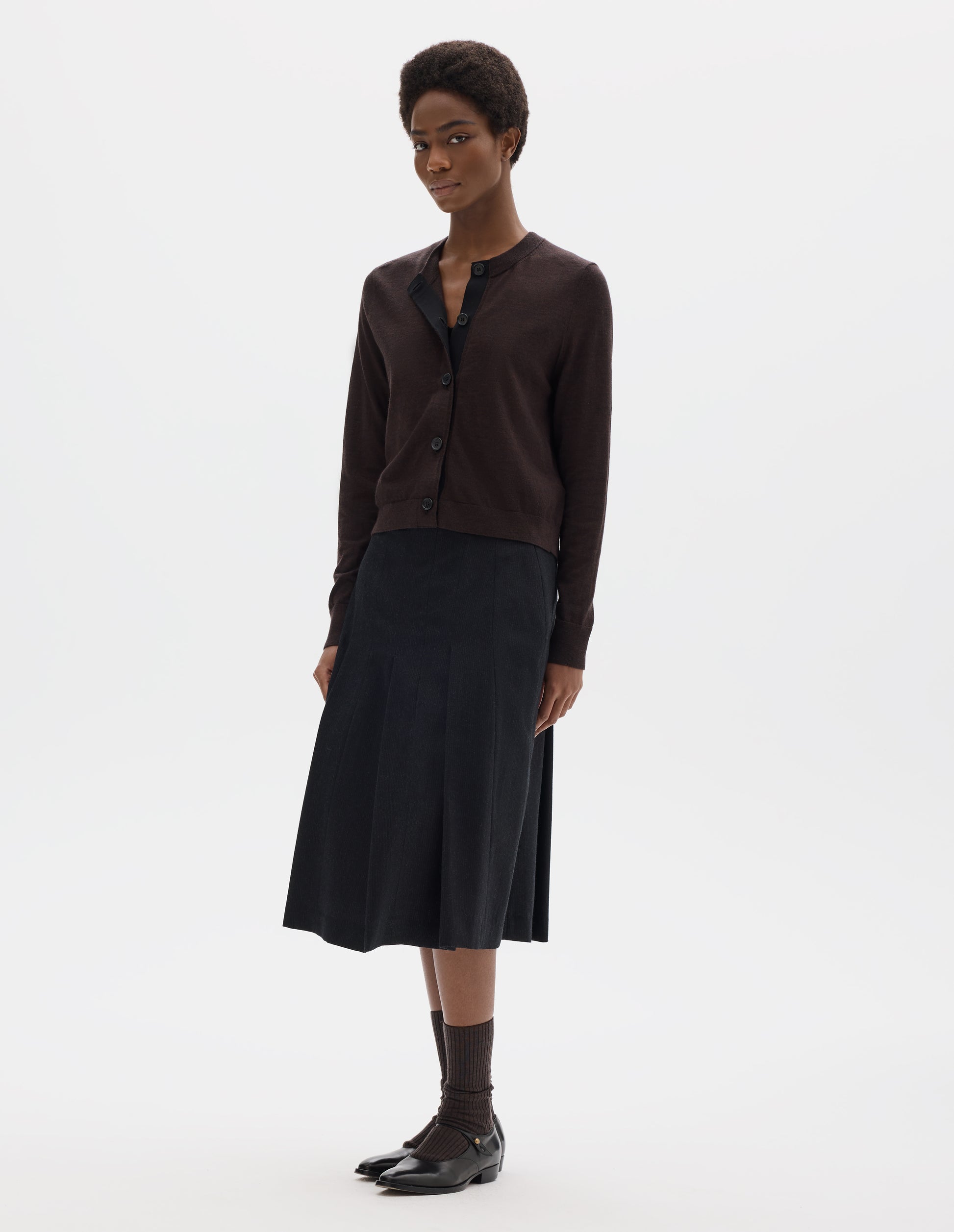 Full body image, front view of model wearing dark brown merino cardigan with fully fashioned sleeves, horn buttons on concealed placket finished with grosgrain, and ribbed neck, cuffs and hem. Styled with black skirt, brown socks and black leather shoes.