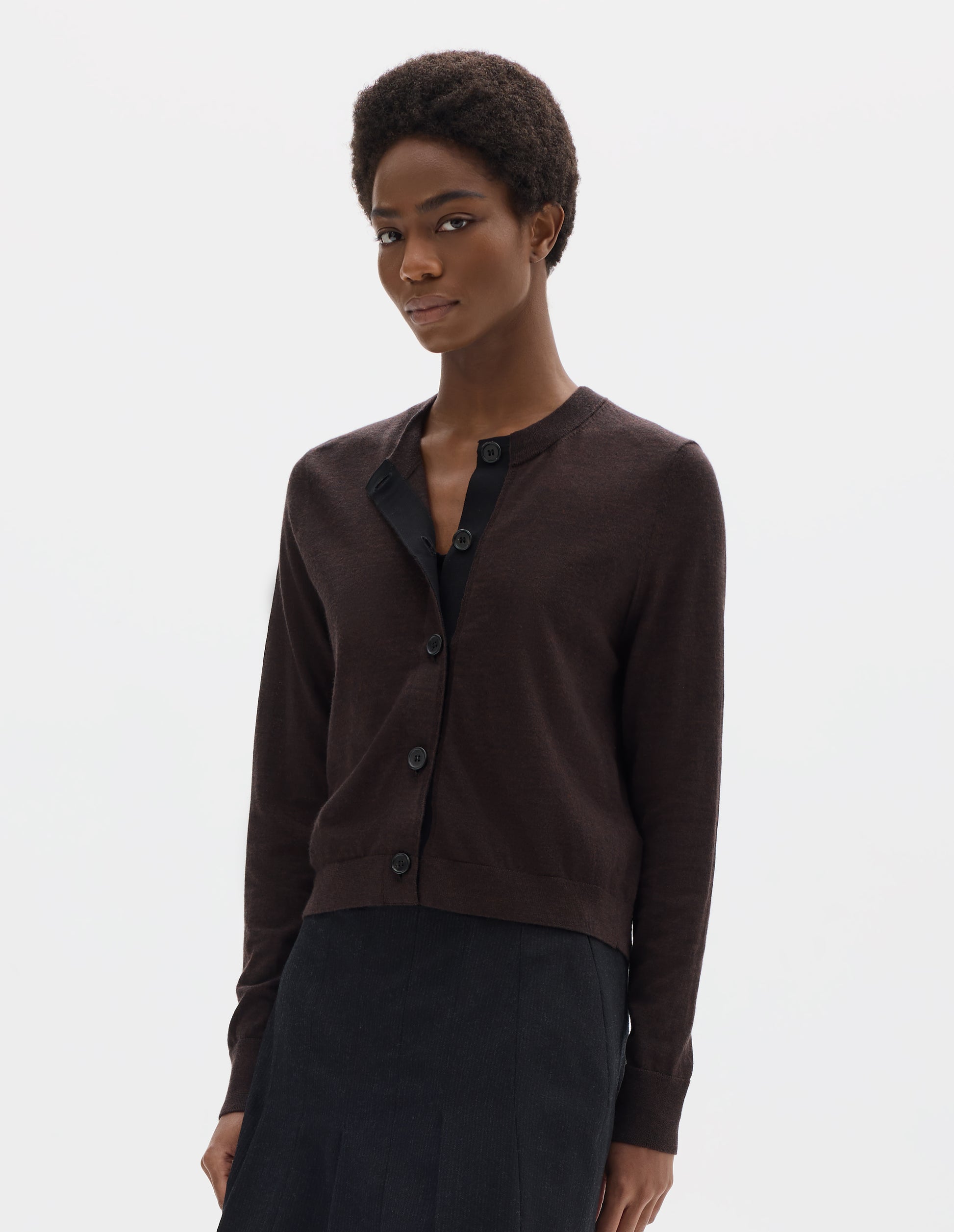 Cropped body image, front view of model wearing dark brown merino cardigan with fully fashioned sleeves, horn buttons on concealed placket finished with grosgrain, and ribbed neck, cuffs and hem. Styled with black skirt.