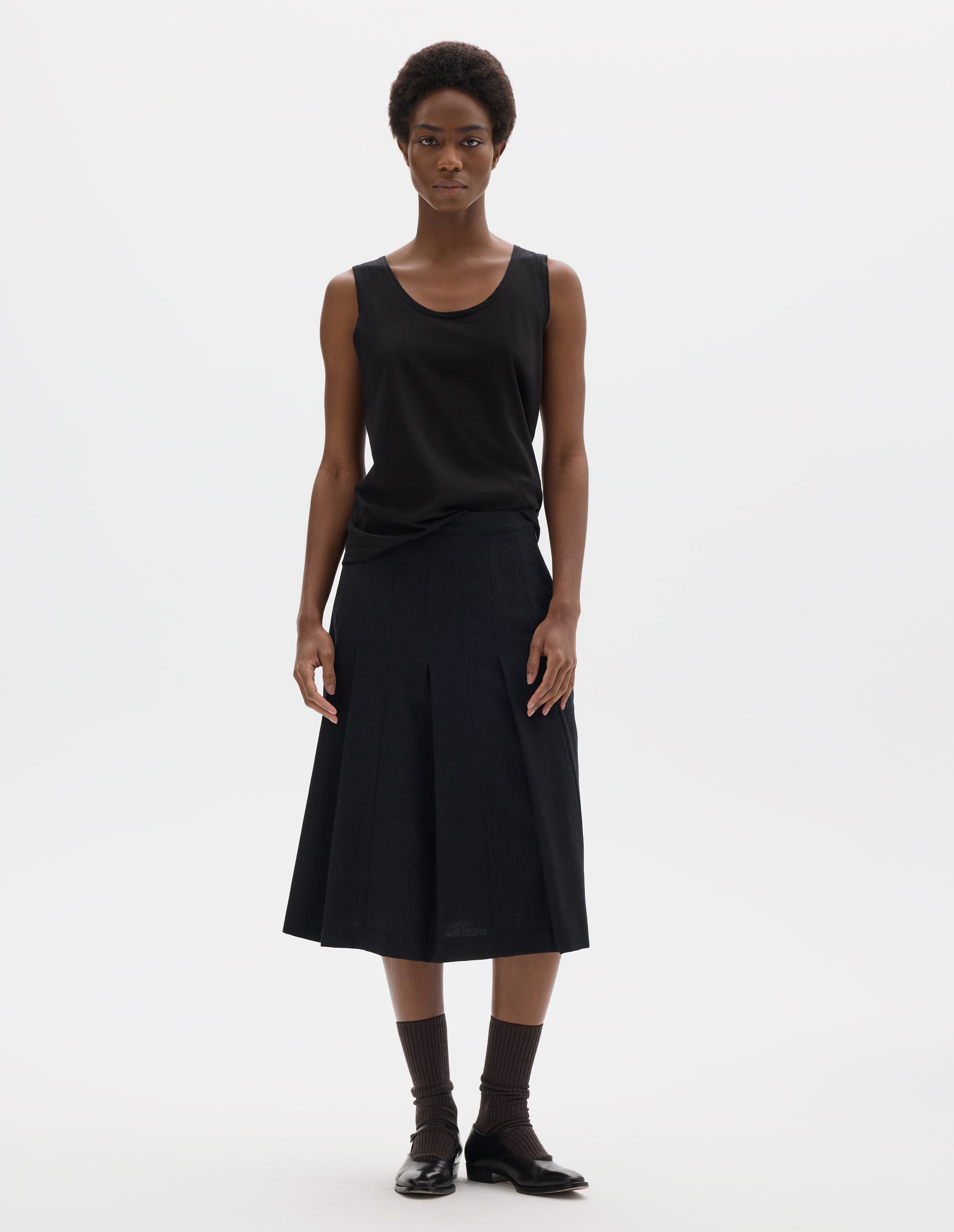 Full body image, front view of model wearing black sleeveless cotton vest with scoop neck. Styled with dark grey skirt, dark brown socks and black leather shoes.