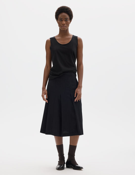 Full body image, front view of model wearing black sleeveless cotton vest with scoop neck. Styled with dark grey skirt, dark brown socks and black leather shoes.