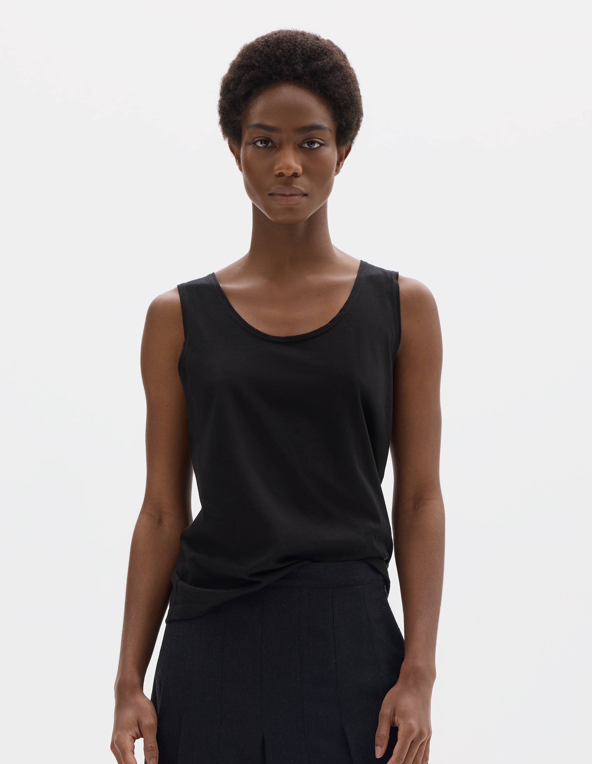 Cropped body image, front view of model wearing black sleeveless cotton vest with scoop neck. Styled with dark grey skirt.